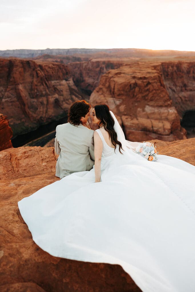 Bride and groom lean in for a kiss as they sit on cliff overlooking Horseshoe Bend, as captured by destination wedding photographer