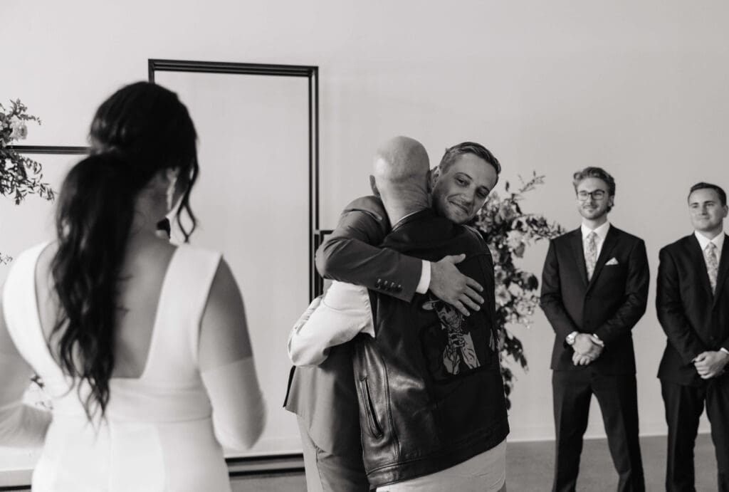 Groom hugs father of the bride while looking at his bride