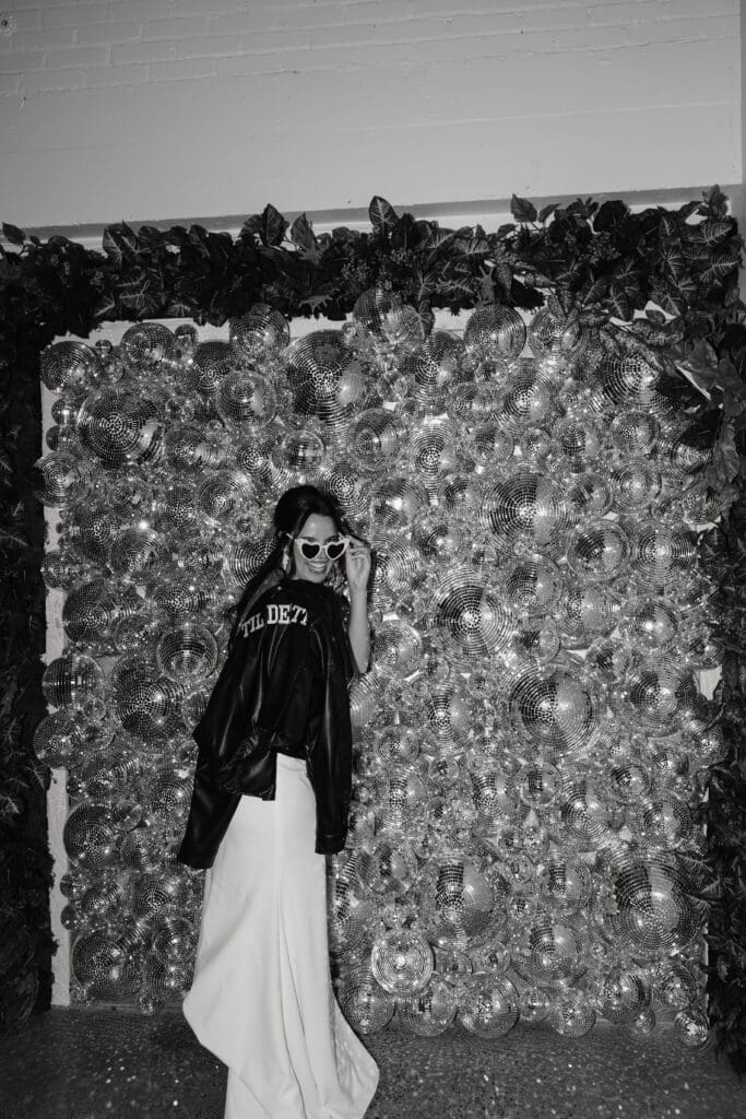 Bride wearing heart sunglasses and leather jacket poses in front of disco ball photo backdrop
