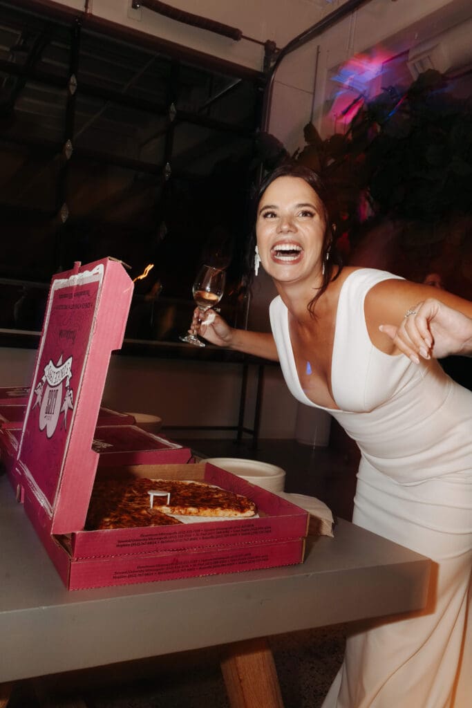 Bride holds glass of wine and digs into huge box of pizza