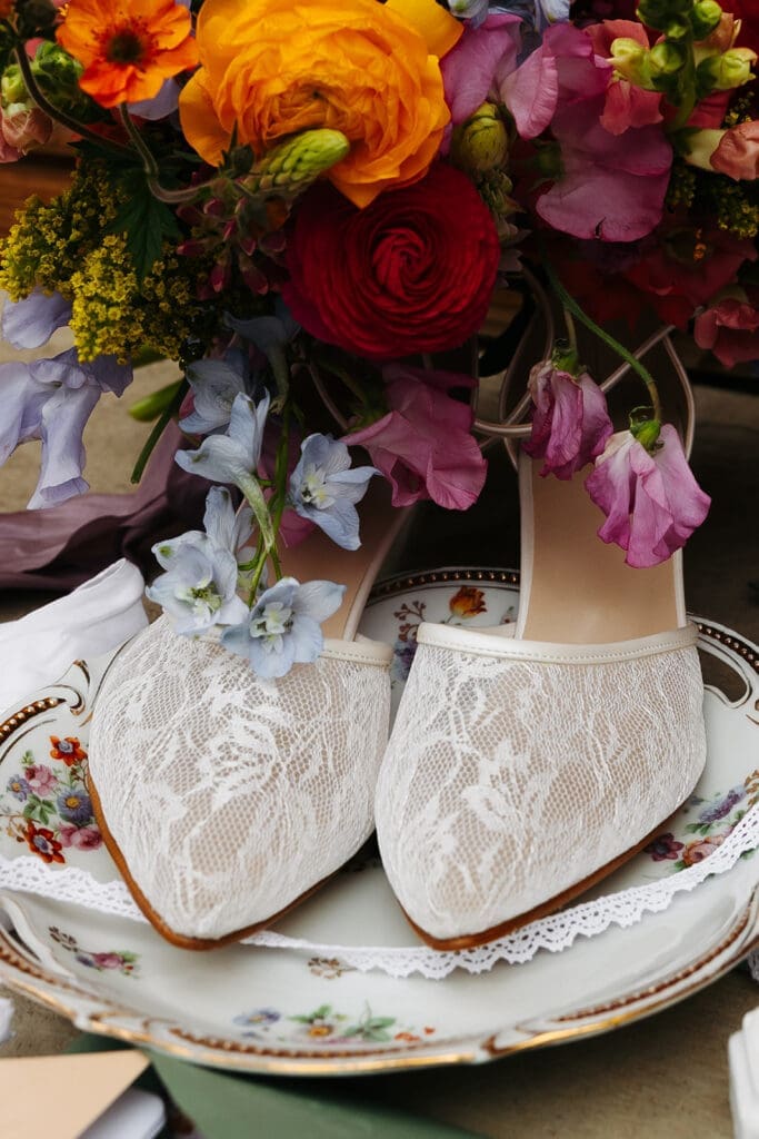 Lace bridal shoes styled on vintage china beneath a lush bouquet of bright seasonal flowers for an editorial wedding flat lay.