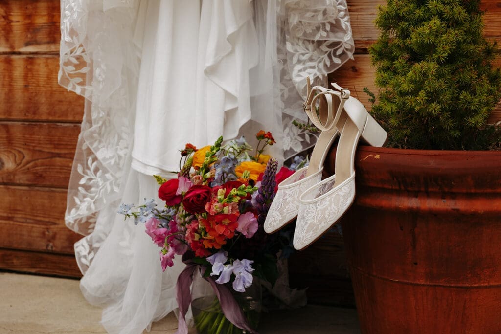 Bridal shoes, colorful bouquet, and wedding dress styled together outside a rustic venue, highlighting personal wedding details and florals.