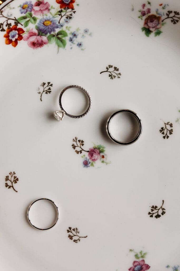 Wedding bands and engagement ring arranged across a delicate floral porcelain plate in a minimalist wedding detail photo.