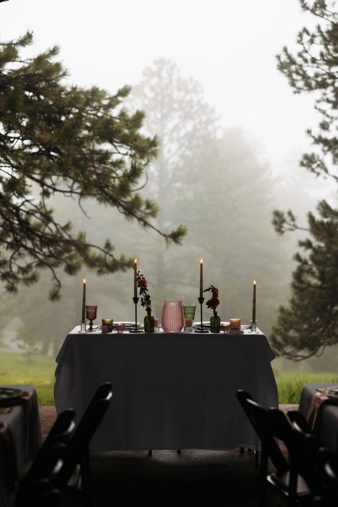 Reception table set beneath towering pine trees on a misty morning, featuring candlelight, florals, and moody woodland wedding styling.