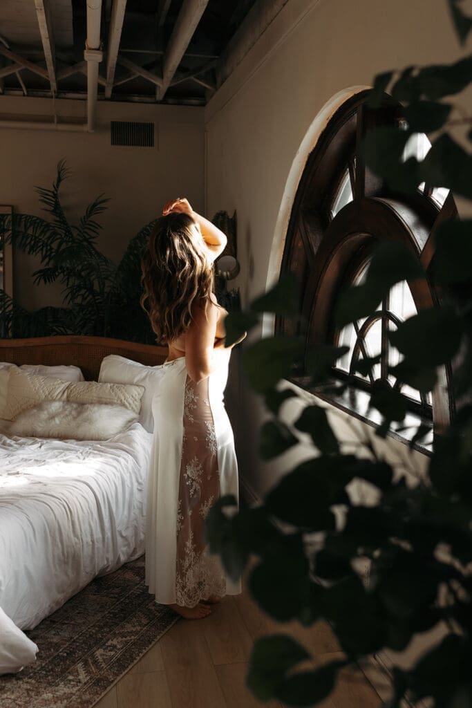 Bride stands in a lace robe near a bedroom window during a bridal boudoir session filled with soft natural light and maximum romance.