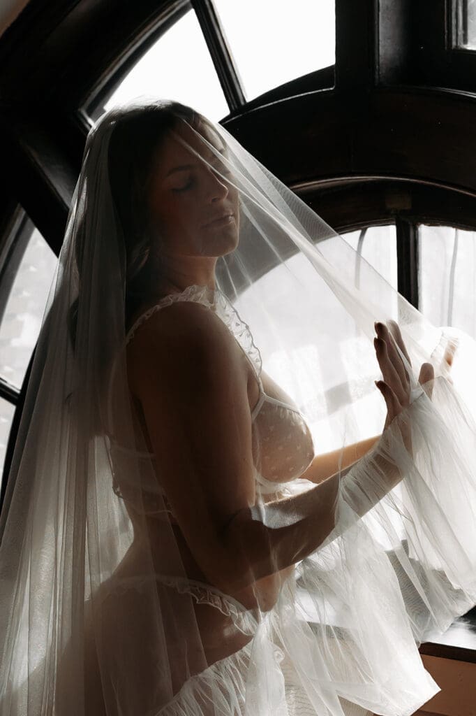 Bride posed beside an arched window beneath a flowing veil during an intimate bridal boudoir session before the wedding day.