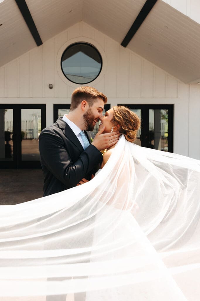 Newlyweds embracing beneath a flowing veil outside the bright white exterior of this modern blank canvas wedding venue.