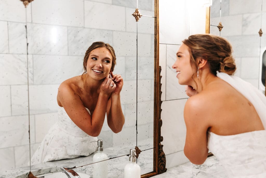 Bride putting on earrings in the bright marble bridal suite at Equestria West, a modern blank canvas wedding venue.
