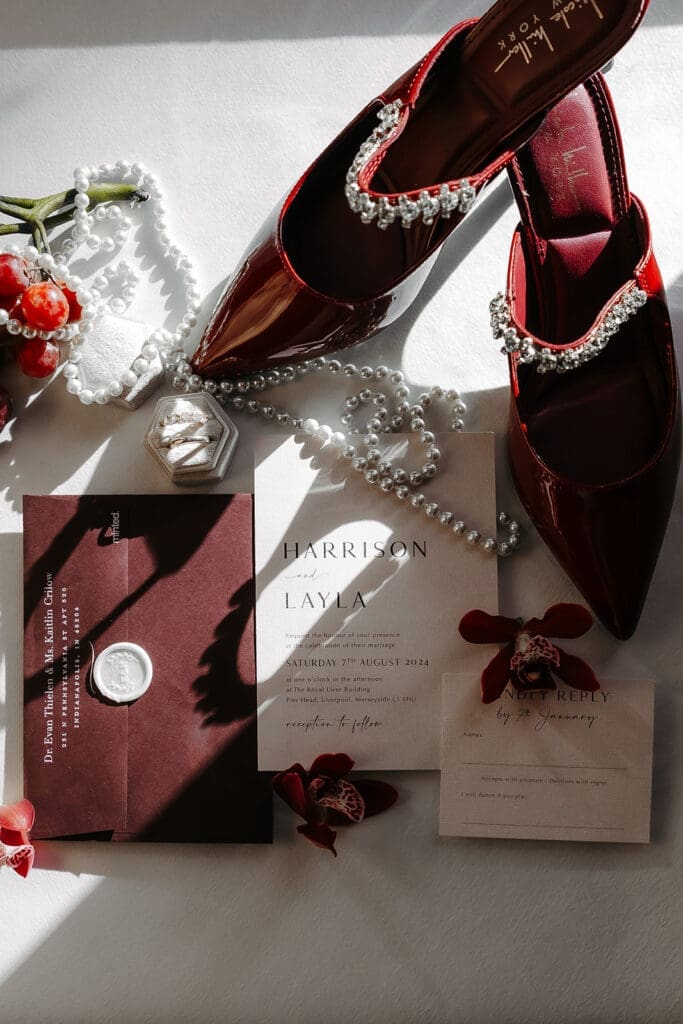 Wedding invitation suite, heirloom pearls, ring box, and ruby-toned bridal shoes arranged in layered natural light.