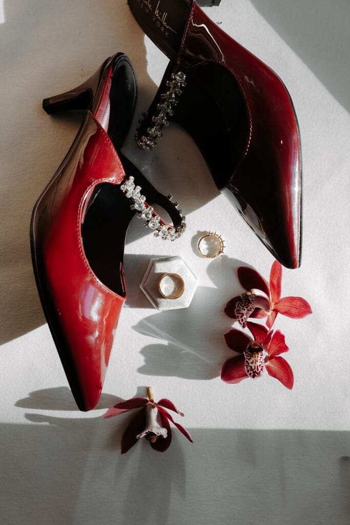 Deep red bridal heels styled with jewelry and orchids in dramatic window light for a fashion-forward wedding flat lay photo.