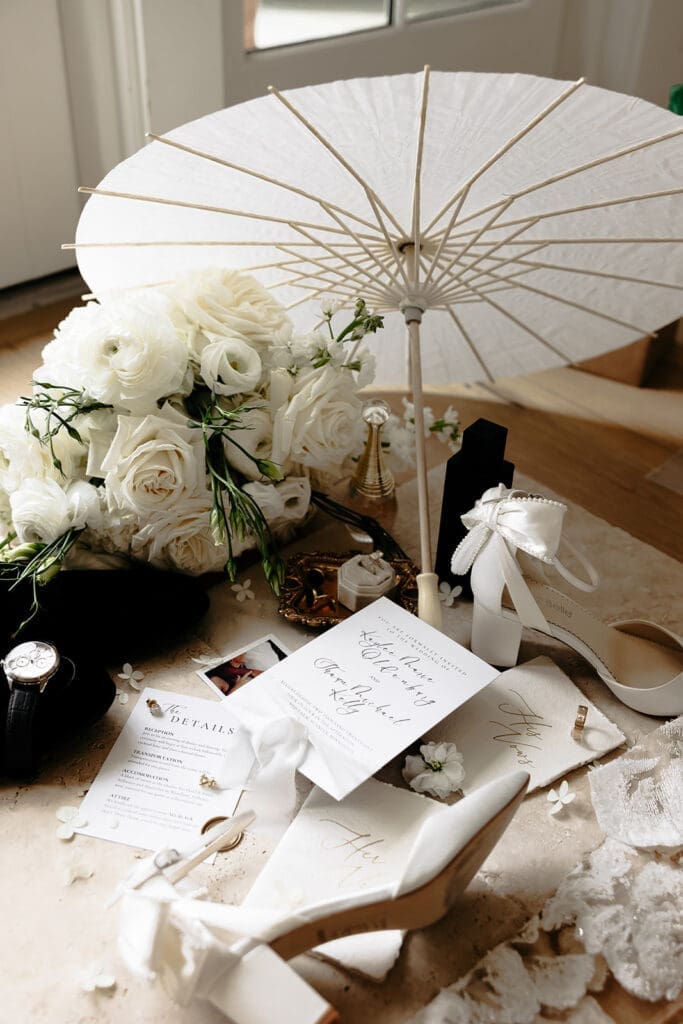 Wedding flat lay featuring bridal shoes, white florals, invitation suite, parasol, jewelry, and meaningful keepsakes gathered from the wedding day.