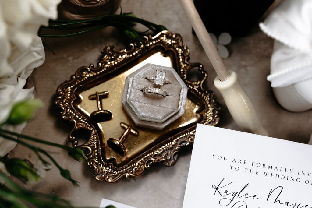 Wedding flat lay photo featuring diamond wedding rings, gold cufflinks, invitation stationery, florals, and vintage brass styling pieces.