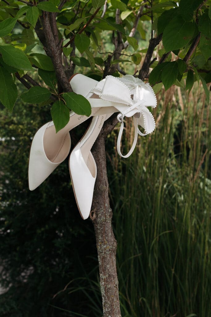 White bridal heels hanging from a tree branch beside tall grasses, photographed as an artistic wedding detail photo.