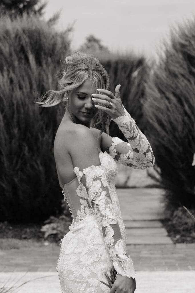 Bride in a floral lace wedding gown surrounded by tall grasses and lush landscaping before the ceremony.
