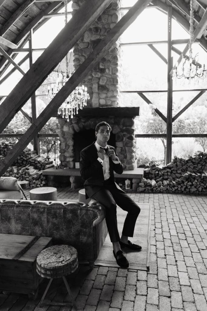 Groom relaxing beside a stone fireplace inside one of the elegant gathering spaces at this estate wedding venue.
