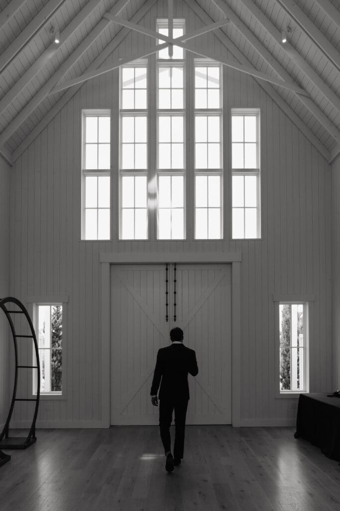 Groom walking through the white barn beneath soaring windows before his first look at Redeemed Farm MN.
