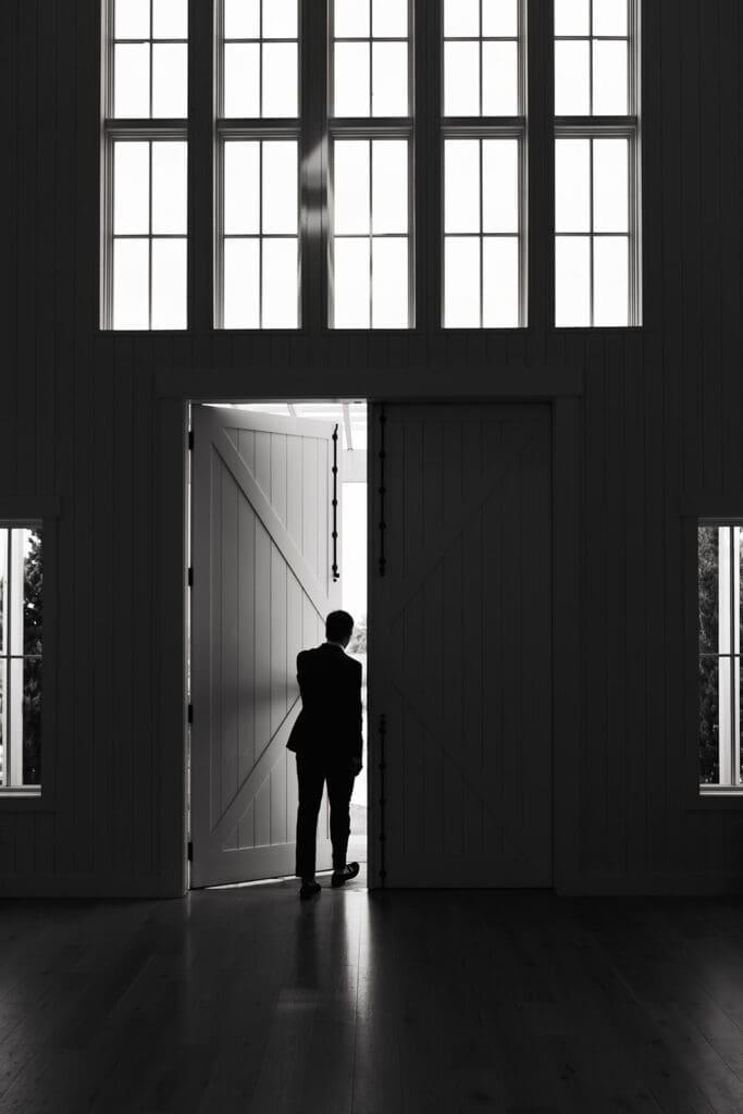Groom stands silhouetted in a doorway waiting for his first look, one of the most emotional wedding photo tips couples can consider for a more relaxed wedding day.