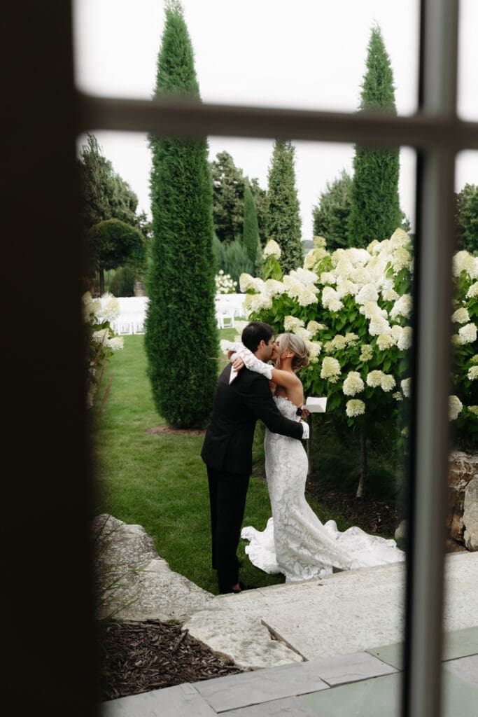 Bride and groom embracing among blooming hydrangeas, photographed through a nearby window for a layered perspective.