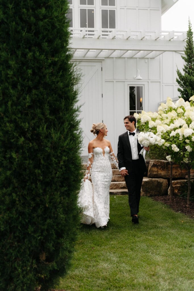 Bride and groom walking through the hydrangea-lined gardens beside the white barn after their ceremony.