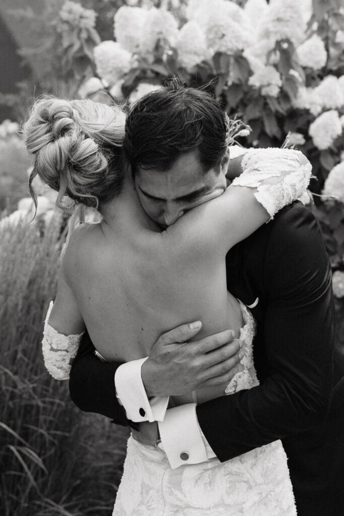 Bride wrapping her arms around her groom among lush white hydrangeas during sunset portraits.