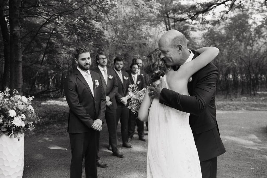 Bride hugs her father before the ceremony while the groom and groomsmen wait nearby during an emotional outdoor wedding, highlighting meaningful moments captured through wedding photography and videography packages.