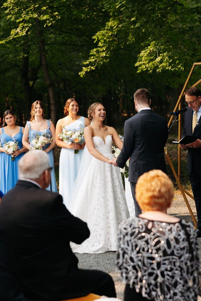 Bride and groom smile during an outdoor wedding ceremony surrounded by their wedding party and guests, creating a moment of maximum romance.