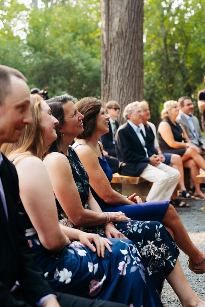 Family and friends smile from the ceremony seats as they watch the couple exchange vows during an outdoor wedding ceremony.