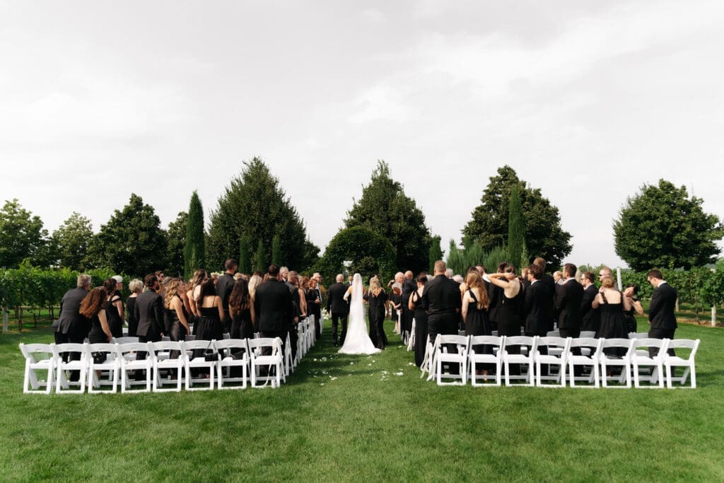 View from the back of the ceremony at vineyard wedding venue in Minnesota as bride makes her way down the aisle towards groom. 