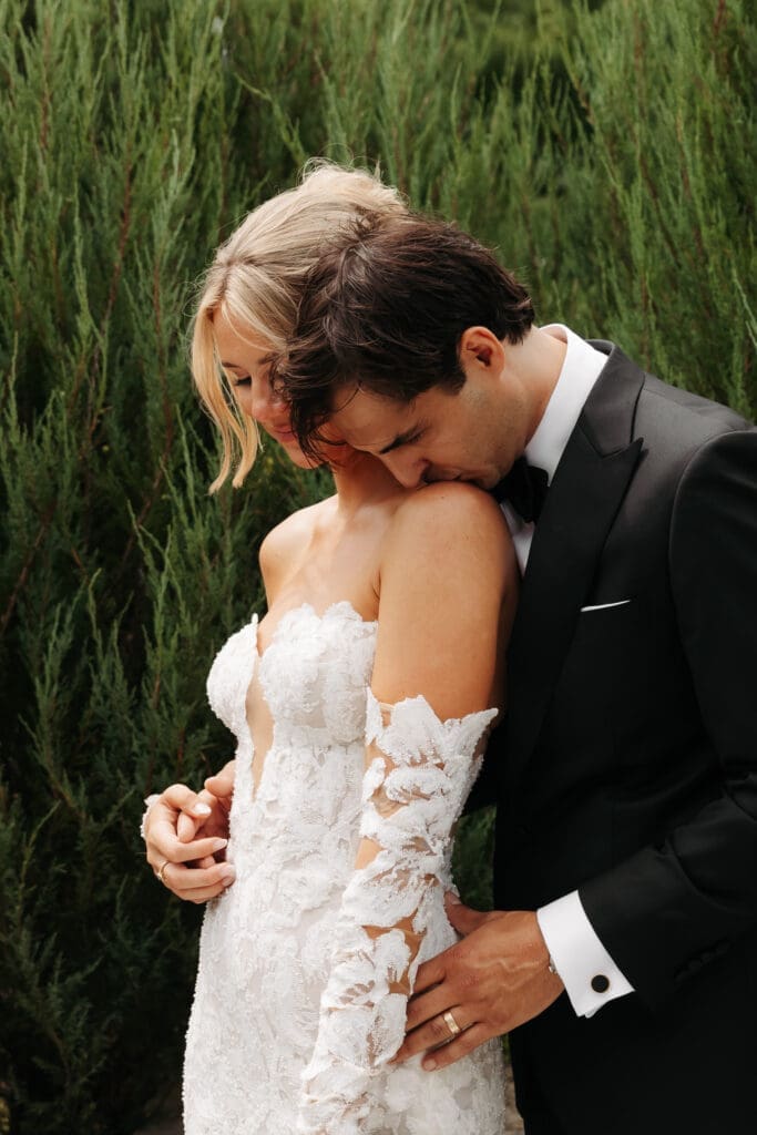 Groom resting his head against his bride's shoulder during portraits surrounded by lush evergreens and garden landscaping at a vineyard wedding venue.