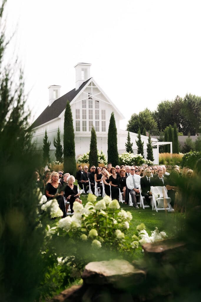 Outdoor wedding ceremony framed through garden florals with guests seated in front of a white chapel, demonstrating creative composition techniques photographers use throughout a wedding day.