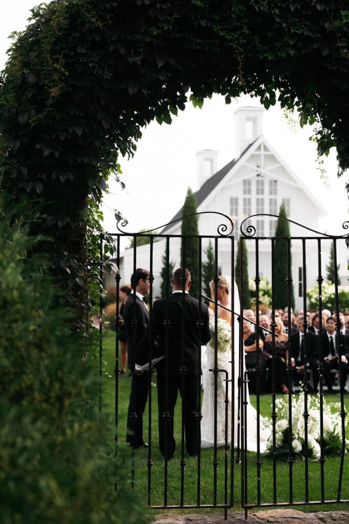 Wedding ceremony photographed through an iron garden gate, showcasing the artistic perspective included in wedding photography and videography packages.