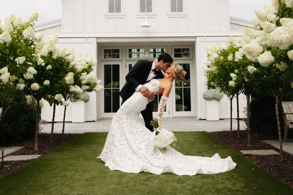 Bride and groom sharing a dip kiss in front of the iconic white barn framed by blooming hydrangea trees.