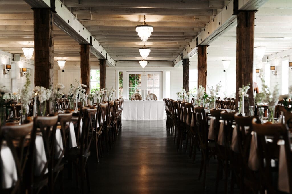 Long reception tables styled with white florals and chandeliers inside the reception hall at an estate wedding venue.