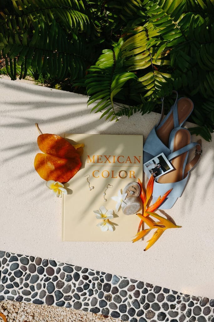 Destination wedding flat lay featuring a travel-inspired book, tropical florals, sandals, and personal keepsakes arranged beside lush greenery.