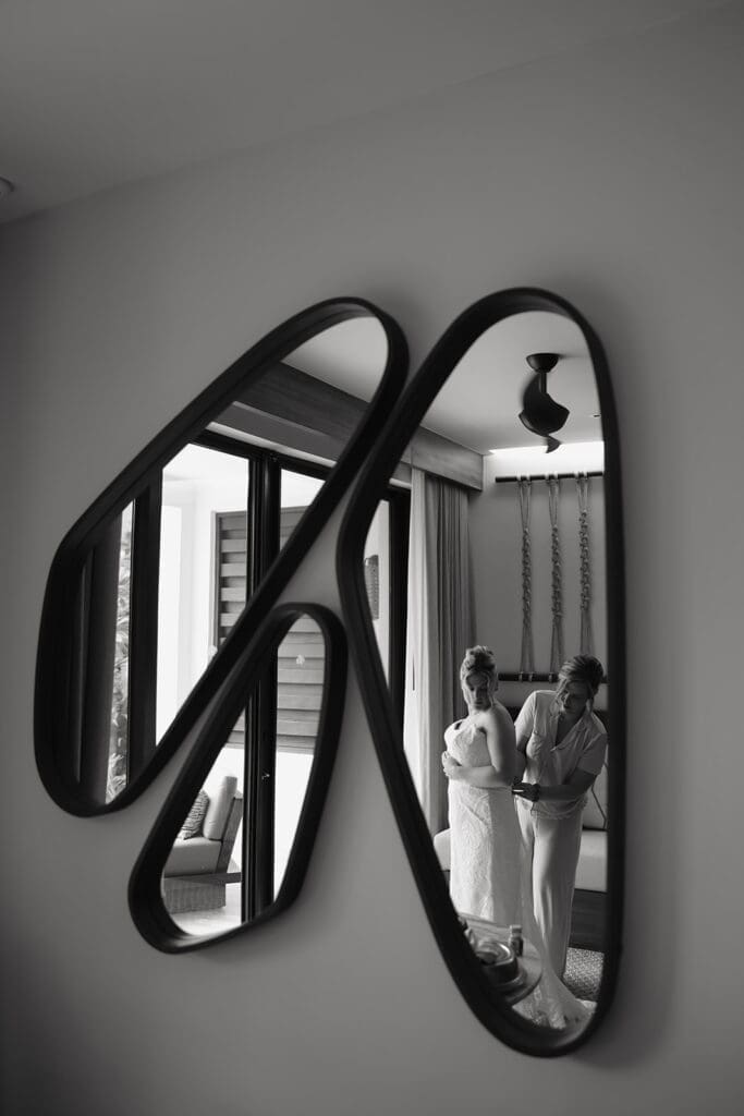 Bride and mother of the bride reflected in a sculptural wall mirror while finishing wedding-day preparations before the ceremony.