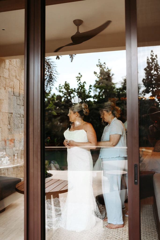 Bride and her mother stand together near a glass door while finishing the final details of getting dressed on the wedding morning.