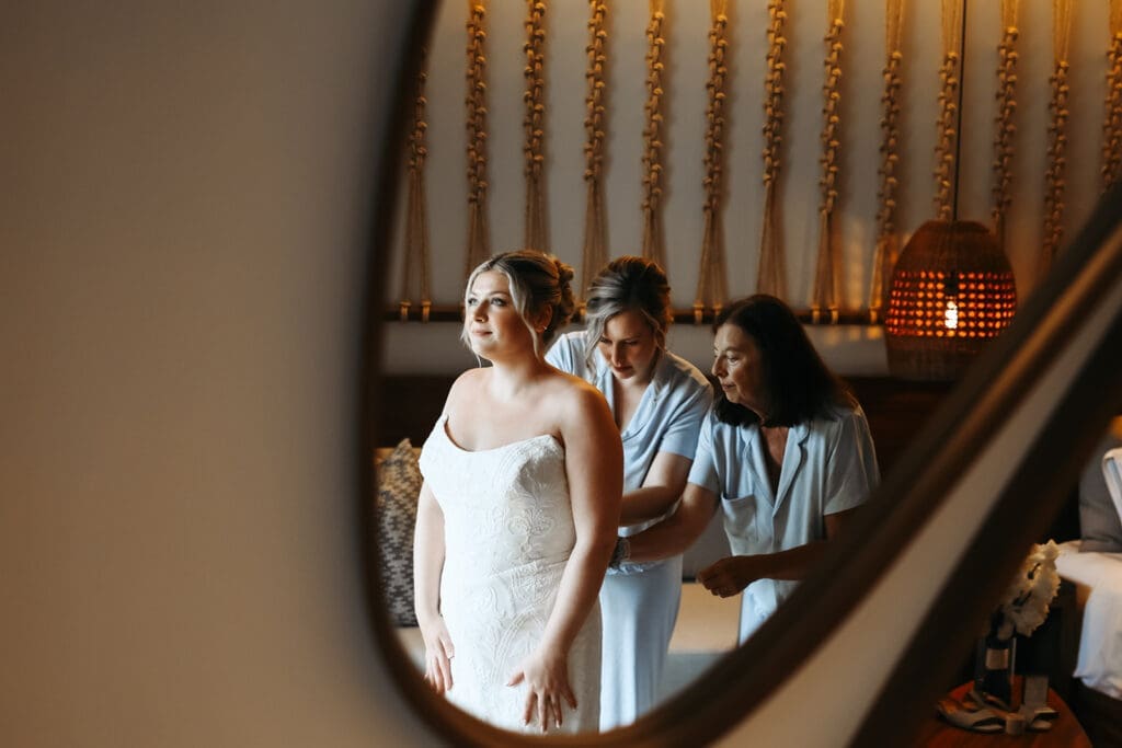 Bride stands in her wedding gown while two women help fasten the dress, photographed through a mirror for an artistic getting-ready moment.