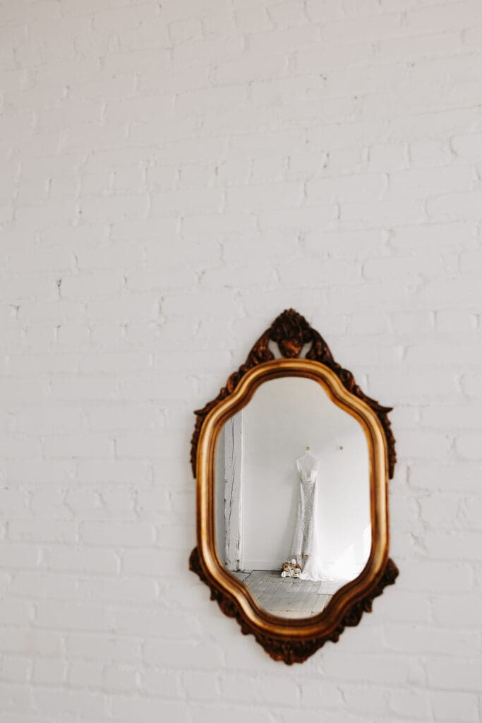 Wedding gown reflected in an ornate vintage mirror against a white brick wall, photographed as an artistic bridal detail.