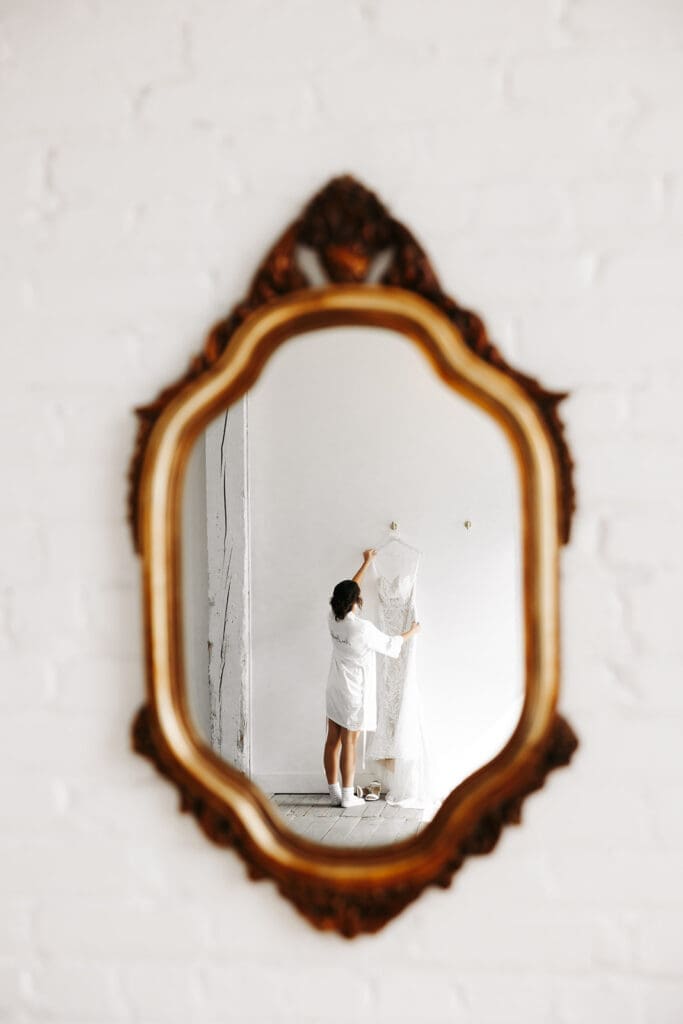 Reflection of a wedding dress hanging in an ornate vintage mirror, creating an artistic bridal preparation photograph.