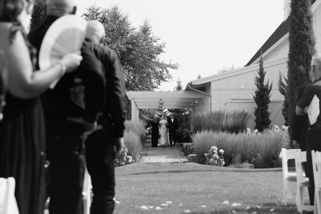 Bride approaching the outdoor ceremony through a garden-lined walkway as guests wait for her arrival at an estate wedding venue.