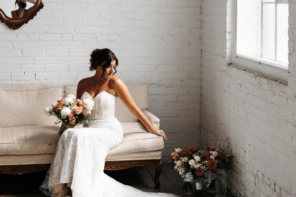 Bride seated on a vintage sofa holding her bouquet during bridal portraits, surrounded by soft natural light and floral arrangements.