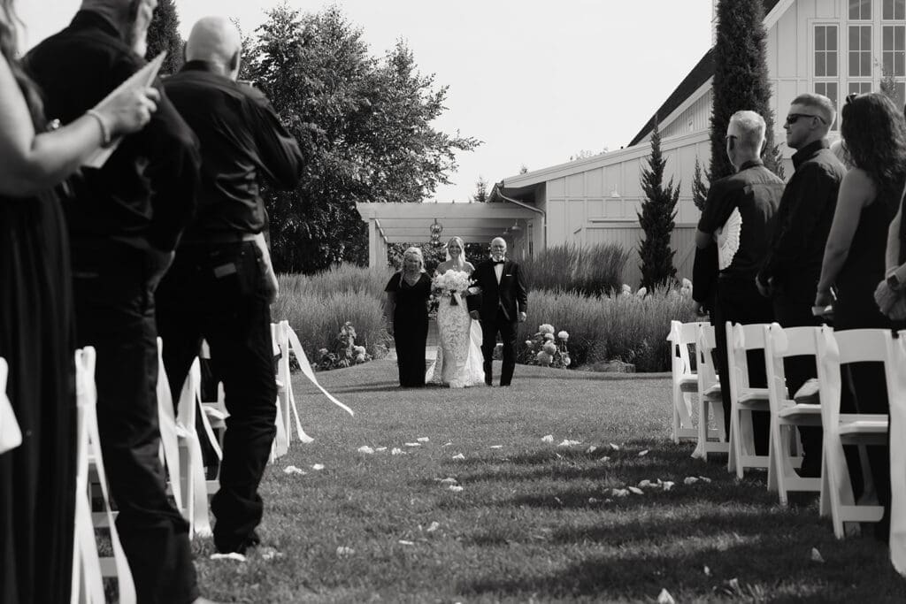 Bride walks down a long outdoor ceremony aisle with her parents as guests look on from both sides, illustrating one of the most valuable wedding photo tips for creating emotional aisle photos.