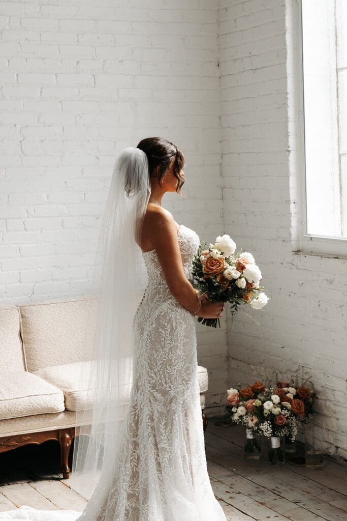 Bride stands beside a bright window holding her bouquet during elegant bridal portraits before the ceremony.