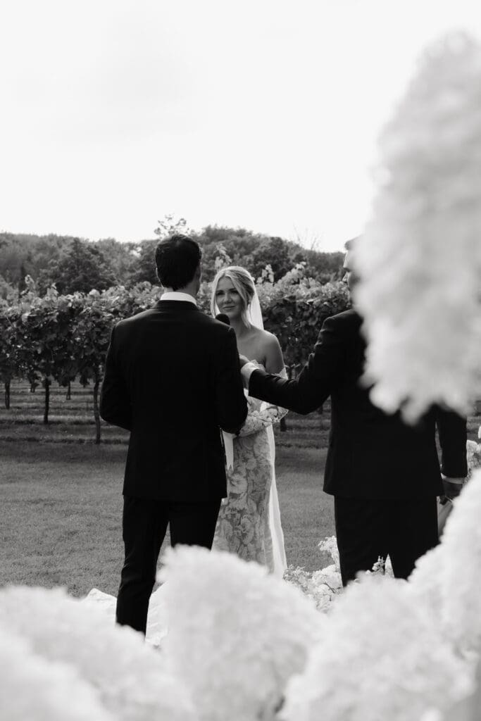 Bride and groom exchanging vows during their outdoor ceremony, photographed through white hydrangea blooms.