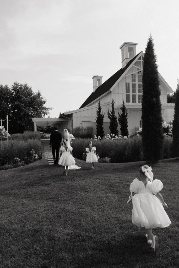 Flower girls racing across the lawn toward the white barn during a summer wedding celebration.