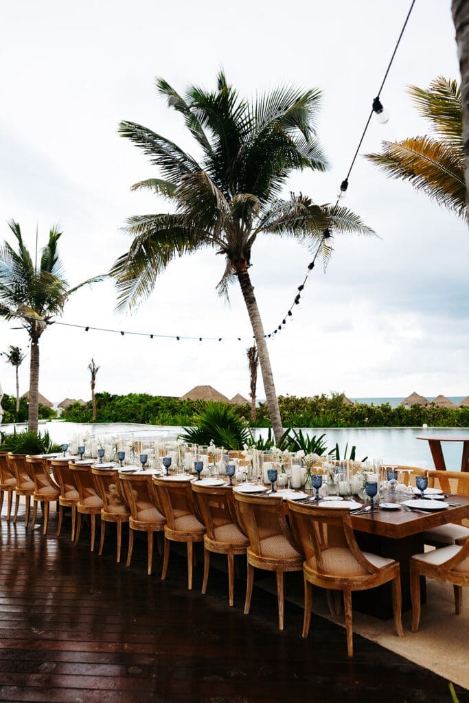 Long waterfront reception table lined with woven chairs, palm trees, and candlelit place settings at a tropical destination wedding.
