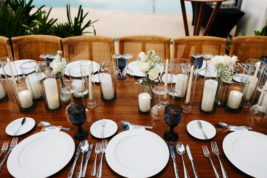 Modern reception tablescape with blue goblets, layered candlelight, and white floral arrangements styled along a warm wood dining table.