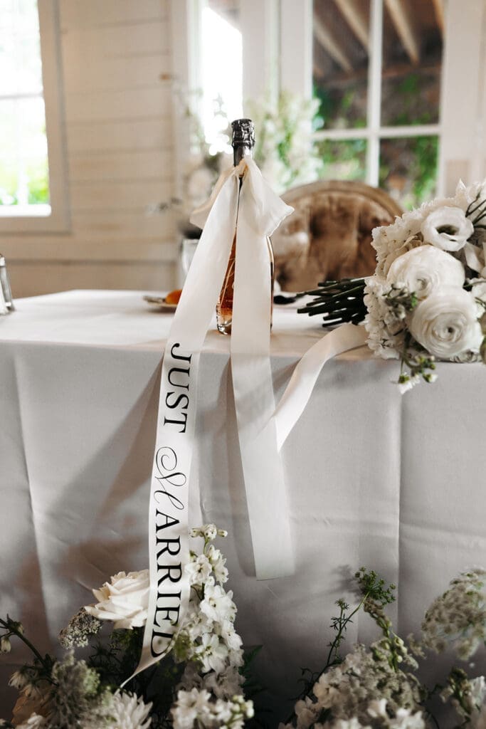 Champagne bottle dressed with a custom "Just Married" ribbon surrounded by white florals and reception decor.