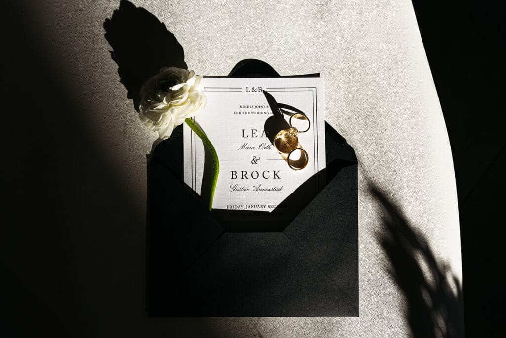 Modern wedding flat lay featuring a black envelope, invitation suite, gold rings, and white ranunculus arranged in dramatic window light.