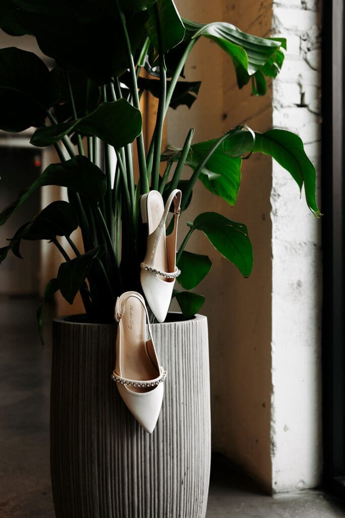 Bridal heels suspended from a large monstera plant inside an industrial wedding venue, photographed as an artistic bridal detail.